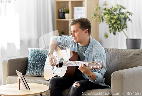 Image of young man with tablet pc playing guitar at home