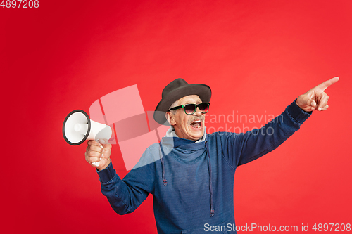 Image of Senior man in stylish eyewear and hat isolated on red background. Tech and joyful elderly lifestyle concept