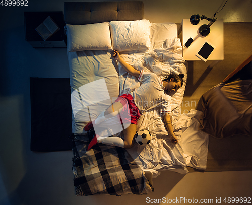 Image of Top view of young professional football, soccer player sleeping at his bedroom in sportwear with ball