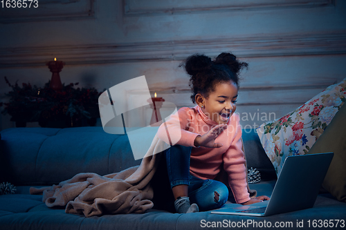 Image of Happy african-american little girl during video call with laptop and home devices, looks delighted and happy