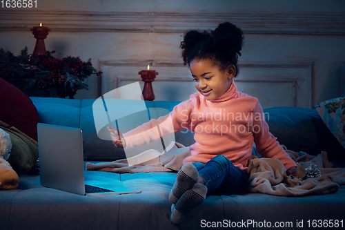 Image of Happy african-american little girl during video call with laptop and home devices, looks delighted and happy