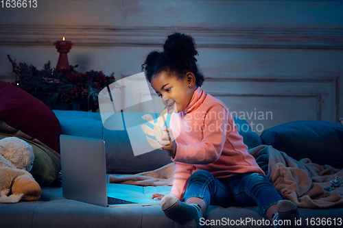 Image of Happy african-american little girl during video call with laptop and home devices, looks delighted and happy