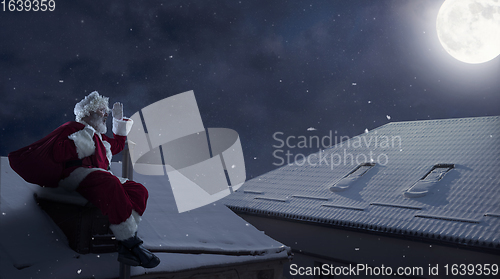 Image of Emotional Santa Claus congratulating with New Year and Christmas, sitting on roof of the house in midnight with full moon