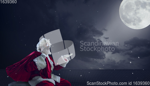 Image of Emotional Santa Claus congratulating with New Year and Christmas, sitting on roof of the house in midnight with full moon