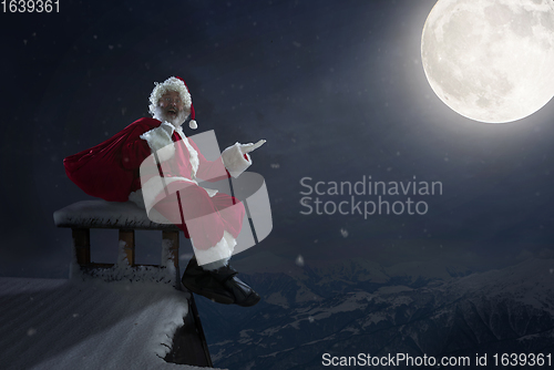 Image of Emotional Santa Claus congratulating with New Year and Christmas, sitting on roof of the house in midnight with full moon