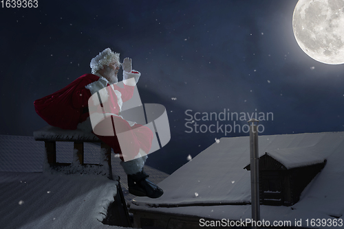 Image of Emotional Santa Claus congratulating with New Year and Christmas, sitting on roof of the house in midnight with full moon