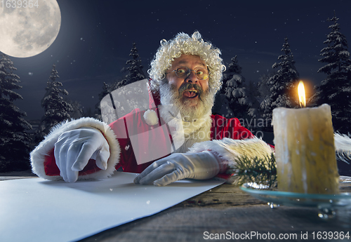 Image of Emotional Santa Claus congratulating with New Year and Christmas, writing a letter, wish list in midnight with candle