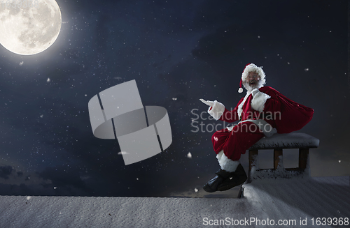 Image of Emotional Santa Claus congratulating with New Year and Christmas, sitting on roof of the house in midnight with full moon