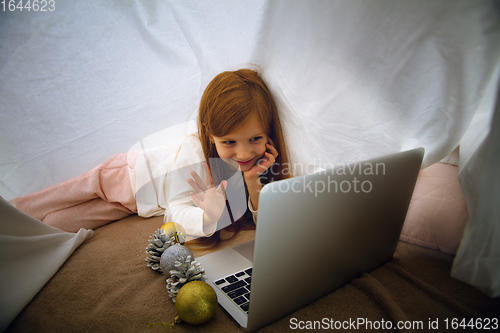 Image of Happy caucasian little girl during video call or messaging with Santa using laptop and home devices, looks delighted and happy