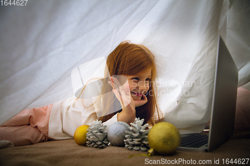 Image of Happy caucasian little girl during video call or messaging with Santa using laptop and home devices, looks delighted and happy