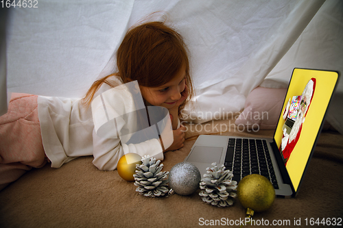 Image of Happy caucasian little girl during video call or messaging with Santa using laptop and home devices, looks delighted and happy