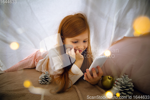 Image of Happy caucasian little girl during video call or messaging with Santa using laptop and home devices, looks delighted and happy