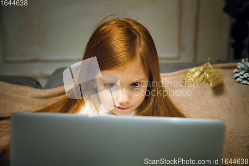 Image of Happy caucasian little girl during video call or messaging with Santa using laptop and home devices, looks delighted and happy