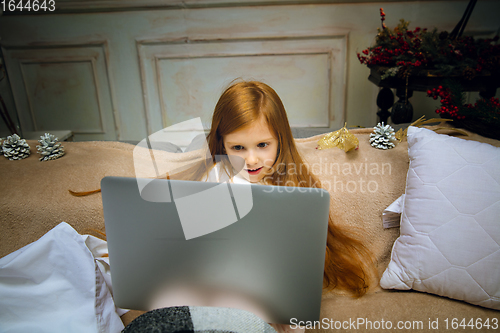 Image of Happy caucasian little girl during video call or messaging with Santa using laptop and home devices, looks delighted and happy