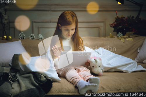 Image of Happy caucasian little girl during video call or messaging with Santa using laptop and home devices, looks delighted and happy