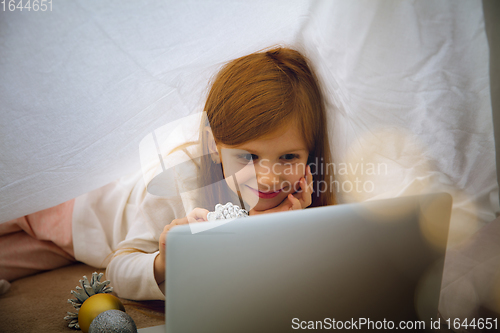 Image of Happy caucasian little girl during video call or messaging with Santa using laptop and home devices, looks delighted and happy