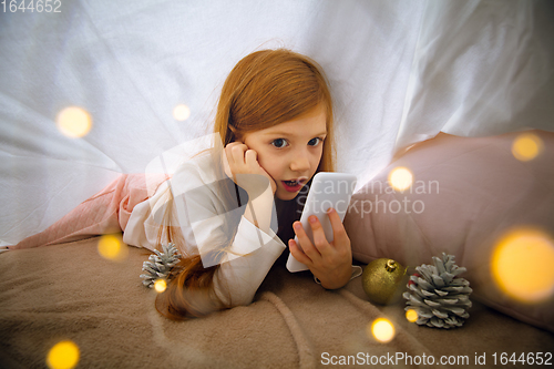 Image of Happy caucasian little girl during video call or messaging with Santa using laptop and home devices, looks delighted and happy