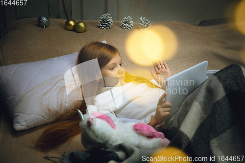 Image of Happy caucasian little girl during video call or messaging with Santa using laptop and home devices, looks delighted and happy