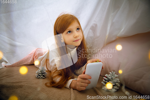 Image of Happy caucasian little girl during video call or messaging with Santa using laptop and home devices, looks delighted and happy