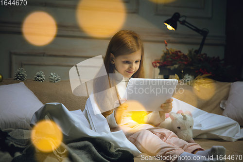 Image of Happy caucasian little girl during video call or messaging with Santa using laptop and home devices, looks delighted and happy