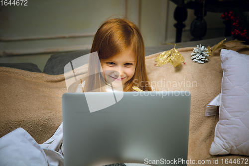 Image of Happy caucasian little girl during video call or messaging with Santa using laptop and home devices, looks delighted and happy