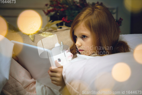 Image of Happy caucasian little girl during video call or messaging with Santa using laptop and home devices, looks delighted and happy