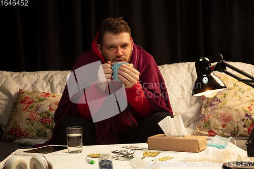 Image of Man wrapped in a plaid looks sick, ill, sneezing and coughing sitting at home indoors