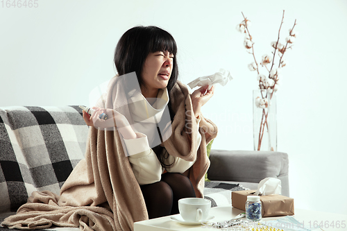 Image of Woman wrapped in a plaid looks sick, ill, sneezing and coughing sitting at home indoors