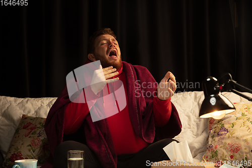 Image of Man wrapped in a plaid looks sick, ill, sneezing and coughing sitting at home indoors