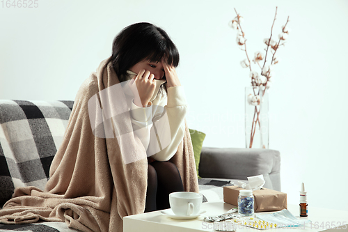 Image of Woman wrapped in a plaid looks sick, ill, sneezing and coughing sitting at home indoors