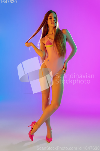 Image of Beautiful seductive girl in fashionable pink swimsuit on bright gradient purple-blue background in neon light