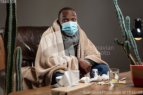 Image of Young man wrapped in a plaid looks sick, ill, sneezing and coughing sitting at home indoors in face mask.