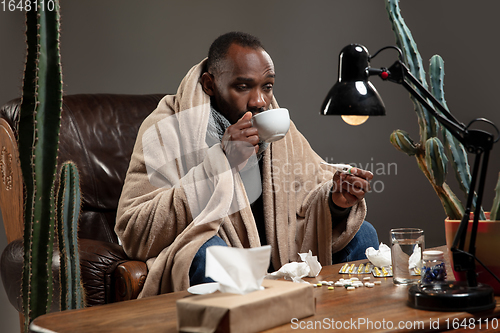 Image of Young man wrapped in a plaid looks sick, ill, sneezing and coughing sitting at home indoors.