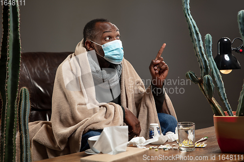 Image of Young man wrapped in a plaid looks sick, ill, sneezing and coughing sitting at home indoors in face mask.