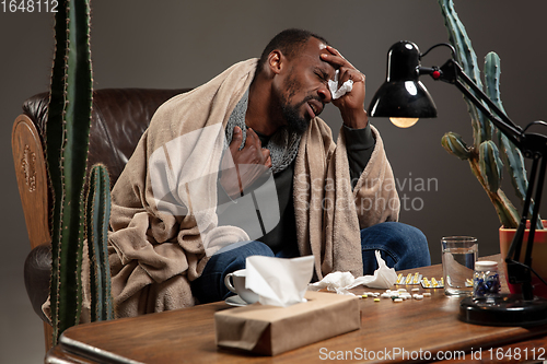Image of Young African-american man wrapped in a plaid looks sick, ill, has terrible headache sitting at home indoors.