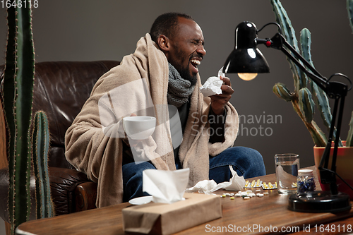 Image of Young man wrapped in a plaid looks sick, ill, sneezing and coughing sitting at home indoors.