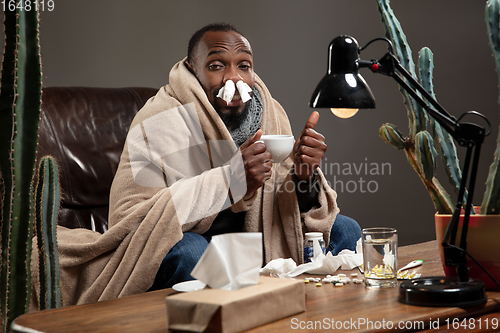 Image of Young African-american man wrapped in a plaid looks sick, ill sitting at home indoors.