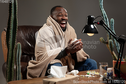Image of Young man wrapped in a plaid looks sick, ill, sneezing and coughing sitting at home indoors.