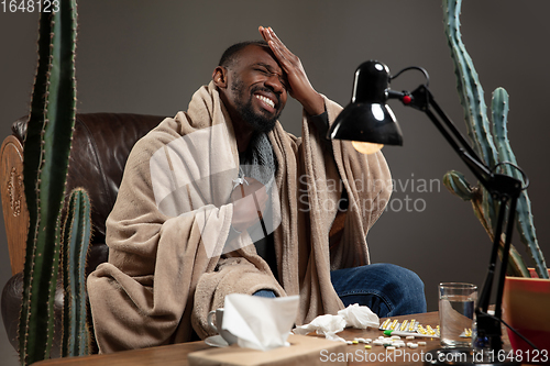 Image of Young African-american man wrapped in a plaid looks sick, ill, has terrible headache sitting at home indoors.