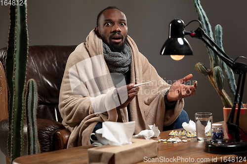 Image of Young African-american man wrapped in a plaid looks sick, ill, sitting at home indoors. Healthcare concept