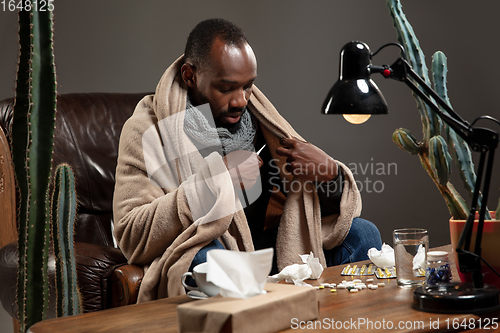 Image of Young African-american man wrapped in a plaid looks sick, ill, sitting at home indoors. Healthcare concept