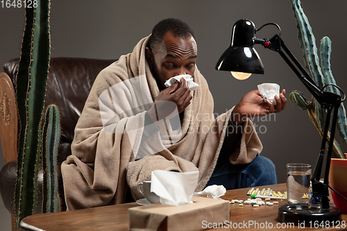 Image of Young man wrapped in a plaid looks sick, ill, sneezing and coughing sitting at home indoors.