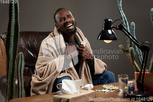 Image of Young African-american man wrapped in a plaid looks sick, ill, sitting at home indoors. Healthcare concept