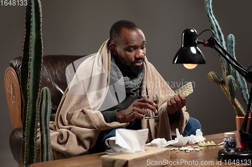 Image of Young man wrapped in a plaid looks sick, ill, sneezing and coughing sitting at home indoors.