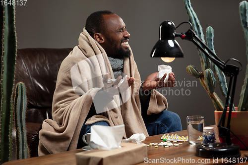 Image of Young man wrapped in a plaid looks sick, ill, sneezing and coughing sitting at home indoors.