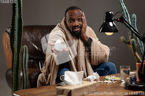 Image of Young African-american man wrapped in a plaid looks sick, ill, has terrible headache sitting at home indoors.