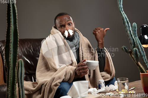 Image of Young African-american man wrapped in a plaid looks sick, ill sitting at home indoors.