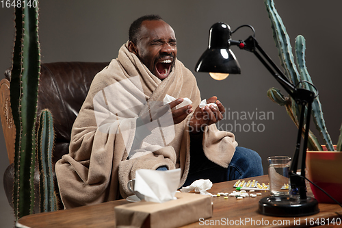 Image of Young man wrapped in a plaid looks sick, ill, sneezing and coughing sitting at home indoors.