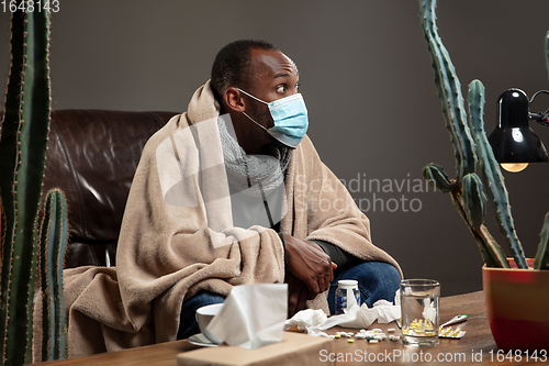 Image of Young man wrapped in a plaid looks sick, ill, sneezing and coughing sitting at home indoors in face mask.