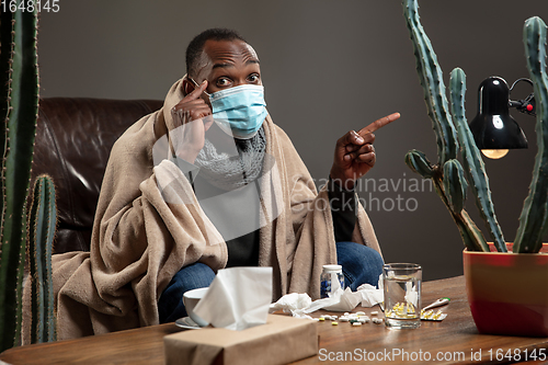 Image of Young man wrapped in a plaid looks sick, ill, sneezing and coughing sitting at home indoors in face mask.